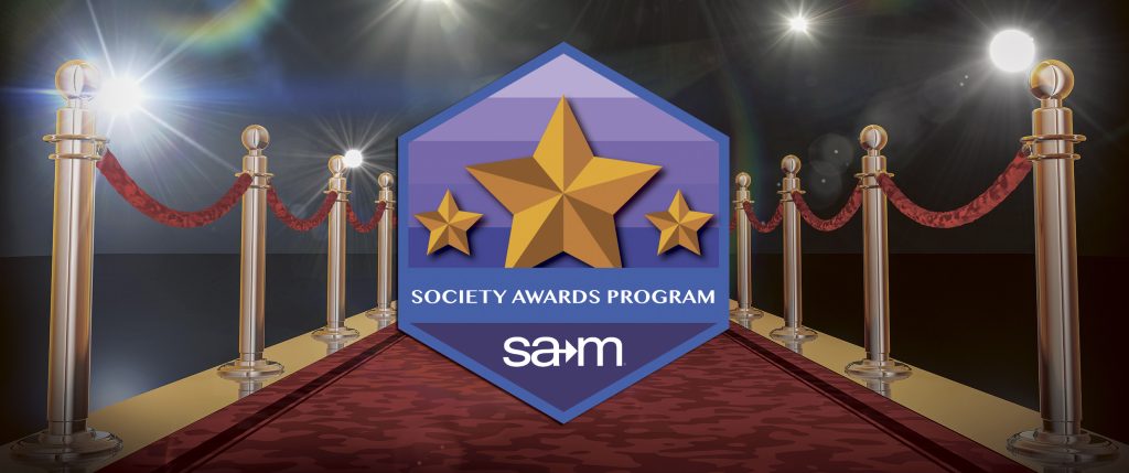 Red carpet event with gold stanchions and velvet ropes leading to a spotlight-lit stage. In the center, a large badge displays three gold stars and the text “Society Awards Program” with the SAM logo beneath it, representing the Society for Advancement of Management’s awards program.