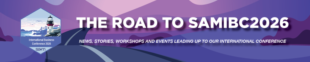 A stylized purple and pink landscape with a winding road leading toward distant hills under a pastel sky, featuring a hexagonal badge on the left showing a lighthouse by the sea and the text ‘International Business Conference 2026 SAM,’ overlaid by bold white lettering reading ‘The Road to SAMIBC2026’ and a subtitle ‘News, stories, workshops and events leading up to our international conference.
